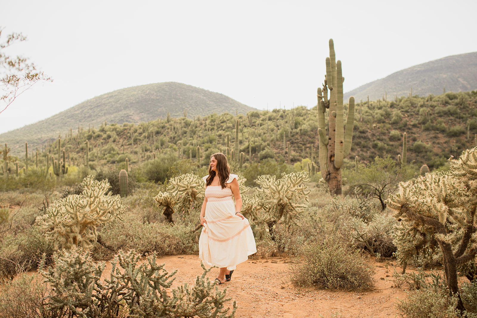 Heather Flynn in a white dress walking through the desert among saguaro cacti and distant mountains